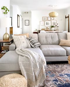 Living Room Sofa Styling featuring a plush couch in a neutral tone. The couch is topped with a textured throw blanket and neutral throw pillows. A patterned area rug anchors the space.