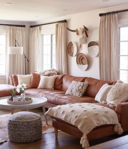 Living Room Sofa Styling showing A spacious living room showcasing a brown leather sectional couch adorned with cream boho throw pillows and a textured throw. A round coffee table in a light wood finish sits in front, complemented by a white cow skull mounted on the wall.